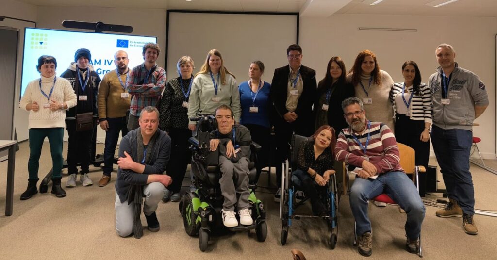 Group photo of TEAM IV project participants and facilitators during an inclusive volunteering training session, with participants standing and seated together in a conference room, including people with disabilities and support staff.