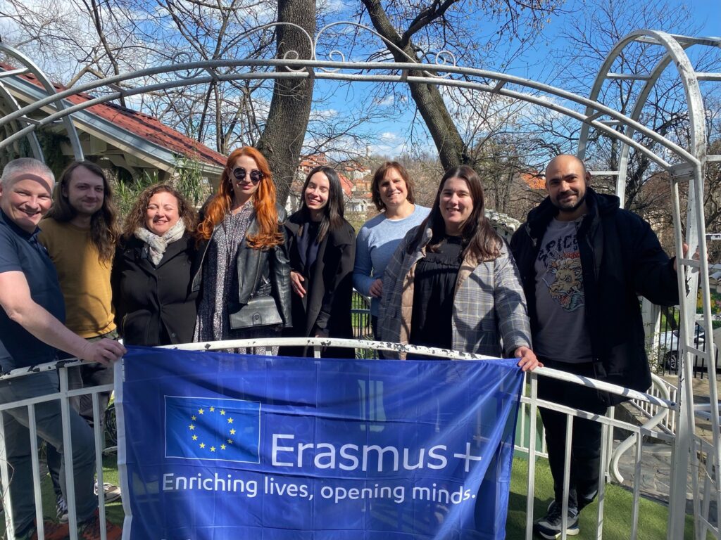 Group photo of TEAM IV project partners holding an Erasmus+ flag outdoors during the international project meeting, standing together in a garden setting on a sunny day.